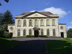 Two story symmetrical building with two tiers of seven windows. Leading up is a driveway with grass and shrubs on either side.