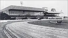 Picture of Rosecroft's track and old grandstand. A horse is racing on the track. To the right, there are several rows of bleachers with a metal tent above for protection.