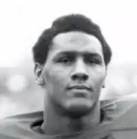 Tight headshot of a dark skinned man with a slightly long afro lookin into the camera. His football shoulder pads are slightly visible.