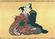 A shunga-style scroll on skill. The background is yellow, and a partially shaved, middle aged men is sitting in green and black robes. Next to him and halfway on his knees is an adolescent boy in an ornate red dress. The two are kissing on the lips with eyes closed; the older man is holding the youth's chin, and the boy's bare feet are exposed.