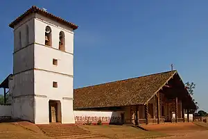 A church and stone bell-tower in three-quarter view. The whitish facade of the church is decorated with motives painted in orange. A wooden cross is positioned at the top of the roof.