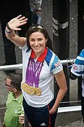 A Caucasian woman with brunette hair waves to a crowd. She is wearing a white polo shirt with the Union Jack flag on the left shoulder. Around her neck hang four Olympic gold medals.