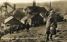 A young girl holds her younger sister on a hillside overlooking the Universal Colliery; a crowd, also waiting for news, are a little further down the hill.