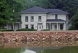 A white house amid tall trees with gently pointed black roofs behind a walkway on a shoreline stabilized with rock. It reflects on water in the foreground.