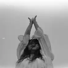 A grayscale photo of a woman dressed in white, raising her hands up toward the sky.