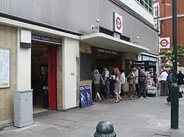 A modern looking building with marble coloured tiled wall at ground level with grey cladding above. A small concrete canopy is over the larger of two entrances with blue signs reading "SLOANE SQUARE STATION", people are entering and exiting through the larger entrance.