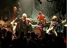  A band consisting of three men and a woman perform onstage in a crowded club in front of a large black sign with "Snowed Out" written in white. The woman stands in the center and leans forward while singing into a microphone. Two guitarists flank her, and the drummer plays behind her.