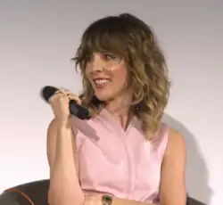 Sophia Di Martino wearing a pink sleeveless top, sitting in an armchair, holding a microphone, grinning, and looking right of camera