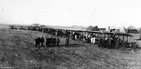 A slightly side-on front view of a line of early aircraft on a flat piece of land. People are standing with the aircraft, with a group standing in front of the machines. Several tent-like structures can be seen in the background.