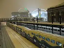 The last commuter train at a train station with a brightly lit stadium nearby. The stadium's roof supports are colored with green and red lights for the Christmas season.