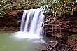 A waterfall into a calm pool along a stream, surrounded by forests.