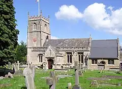 Yellow stone building, with porch with triangular roof in front. Short square tower with battlements topped by flag and flag pole. Gray gravestones in the foreground