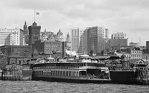 The Whitehall Terminal, a ferry terminal, is in the foreground between skyscrapers behind it and a harbor in front of it.