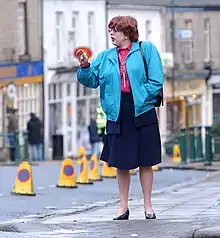 A photograph of a person wearing a light blue jacket, a pink shirt, a navy skirt, navy high-heeled shoes, and glasses while standing on a sidewalk and looking to the left