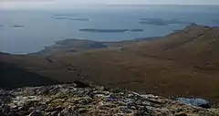 A view from a high rocky eminence with brown moorland below and a vista of small brown islands scattered in the sea beyond. A low bank of fog obscures the horizon.