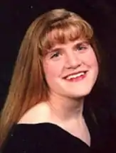 A young white woman with straight reddish-tinted blonde hair that runs down below her shoulders over a black top smiles and looks at the camera.