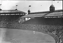 The edge of a cricket field, in front of covered stands crowded with spectators.