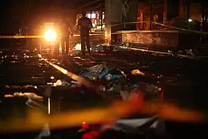 Three police officers standing in a cordoned-off area, one carrying a flashlight. Wreckage from the bombing can be seen.