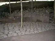 An area of excavation under a corrugated roof supported by wooden poles. The foreground shows an even cobbled pavement running diagonally from left front to right back, where it disappears into a vertical wall of partially excavated rubble and soil.