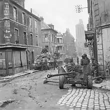 An intersection; an anti-tank gun is covering the crossroads in the foreground and in the street behind are some soldiers and two tanks