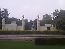 Trees surrounding a white stone obelisk shaped monument, inside a white walled compound, with hedges in the foreground