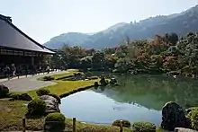 Raked gravel in front of a pond and a wooded hill.