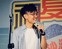 Terence Lam is holding a microphone that says "初秋樂 Band Fun" at the top and "屯門沙灘節2019" at the bottom.