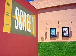 A sign in the foreground reads 'the Screen' while in the background an exterior view of a large, boxy building styled in pseudo-adobe stucco shows off a grassy lawn, a trash can, and lamp-illuminated movie posters