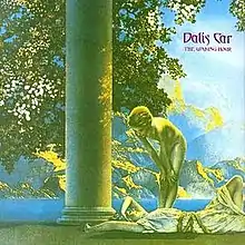 A predominately green, yellow, and blue saturated Ancient Greek-style image of a girl lying down smiling and another girl standing above her, naked, looking down at her. Next to them is a column. The leaves of a tree are visible directly behind the column. In the background is a large mountain, separated by the sea. "Dali's Car", with "The Waking Hour" below it in smaller text, are imprinted in fantasy-style purple text in the upper-right corner.