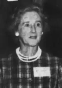 An older white woman wearing pearls and a tartan dress, with a large nametag on her chest