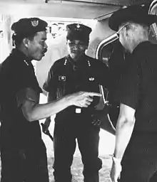 Three Asian men with black hair standing from left to right. The first is in profile, wearing a beret. He has three stars as a Lieutenant General and has a moustache. The next man is facing towards the camera smiling. He is clean shaven and has three stars. A third man on the right has his back to the camera and is wearing a cowboy hat. All are wearing military fatigues.