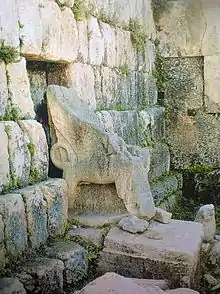 A moss covered stone throne with sphinxes and cloven feet. Behind the throne are moss-covered bare ashlar walls.