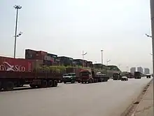 line of heavy trucks going north, background a container yard