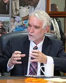 Tim Killeen sits at a wooden table and speaks to Tom Vilsack (unseen) with hands raised