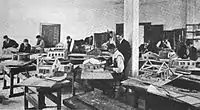 Students building individual houses in a large room with tables