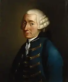 Man in his 50s. He wears a white wig and a blue jacket. He is facing center.