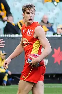 A male athlete with blonde hair wearing a sleeveless jersey and shorts holds football, turning whilst he walks on the grass surface of the playing arena.