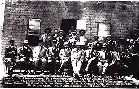 A black-and-white photograph of a group of men standing and sitting in front of a war damaged building. The caption at the bottom lists the names of those present