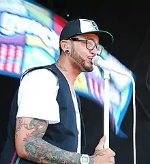 A black male sings into a white microphone whilst wearing a white shirt and black and white baseball cap.