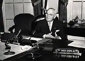 Man in suit sitting behind desk with sign that says "The buck stops here"