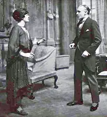 stage scene depicting a youngish white woman and a youngish, clean-shaven white man in morning dress looking at each other