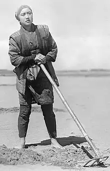 Black and white photograph of Akiko Kazami barefoot with a hoe and standing next to soil that has been dug up