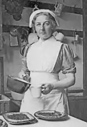 Monochrome photograph of Turner in a cook's apron and hat. She holds a saucepan and a measuring cup. On a table in front of her are several flans, still in their metal dishes