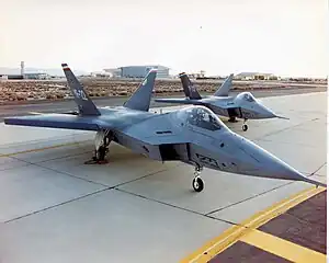 Two jet aircraft with outward-canted vertical stabilizers parked on an angle on ramp.