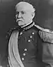 An older white man with Van Dyke mustache and beard wearing an ornate military jacket with fringe at the shoulders, a wide sash across the chest, and a medal hanging from his neck.