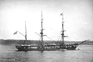 USS Ossipee. The ship lies at anchor in what appears to be an otherwise deserted bay. A low, dark shoreline covered with trees is behind the ship in the middle distance. Ossipee has three masts, but her short, broad smokestack cannot be seen at all because of the dark shoreline behind. The vessel looks to be a natural wood color, excepting two narrow white stripes running the length of the hull, one a little below the main deck and one along the waterline, which is clearly visible. A row of what looks like gunports can be seen just below the upper white line. The ship has a rounded stern and a pronounced clipper bow. The vessel is in the process of lowering a boat, which is hovering just above the sea near the ship's bow.