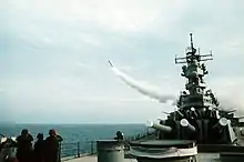 A forward view of a ship with large gun barrels pointing forward. To the left, a missile is flying away from the ship.