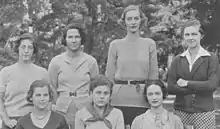 Four women stand behind three seated women, all facing the camera.