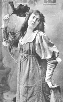 A young white woman, posed with a fan, holding one side of her skirt with her other hand; she is wearing a loose-fitting dress with round baggy upper sleeves and an open neckline; her hair is long and loose