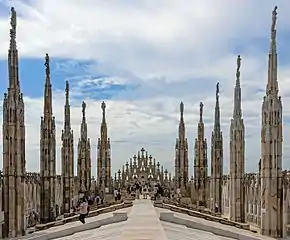 A stone rooftop, sloping down from a level centre to edges where intricately decorated stone spires rise, with statues atop, proceeding toward a rear wall with some more spires. Near the walls, people are gathered. Above is a blue sky largely covered with thin clouds